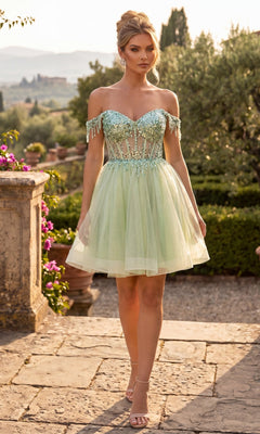 A woman stands outdoors on a stone path surrounded by greenery, wearing the Chicas C356 short homecoming dress in sage—a fit-and-flare, off-the-shoulder style featuring sparkling sequin embellishments and a flattering corset bodice.