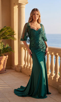 A woman in the Chicas C364 Long Formal Dress in emerald green, featuring sheer floral embroidered sleeves and a subtle leg slit, stands on a balcony overlooking the sea with a potted plant in the background.