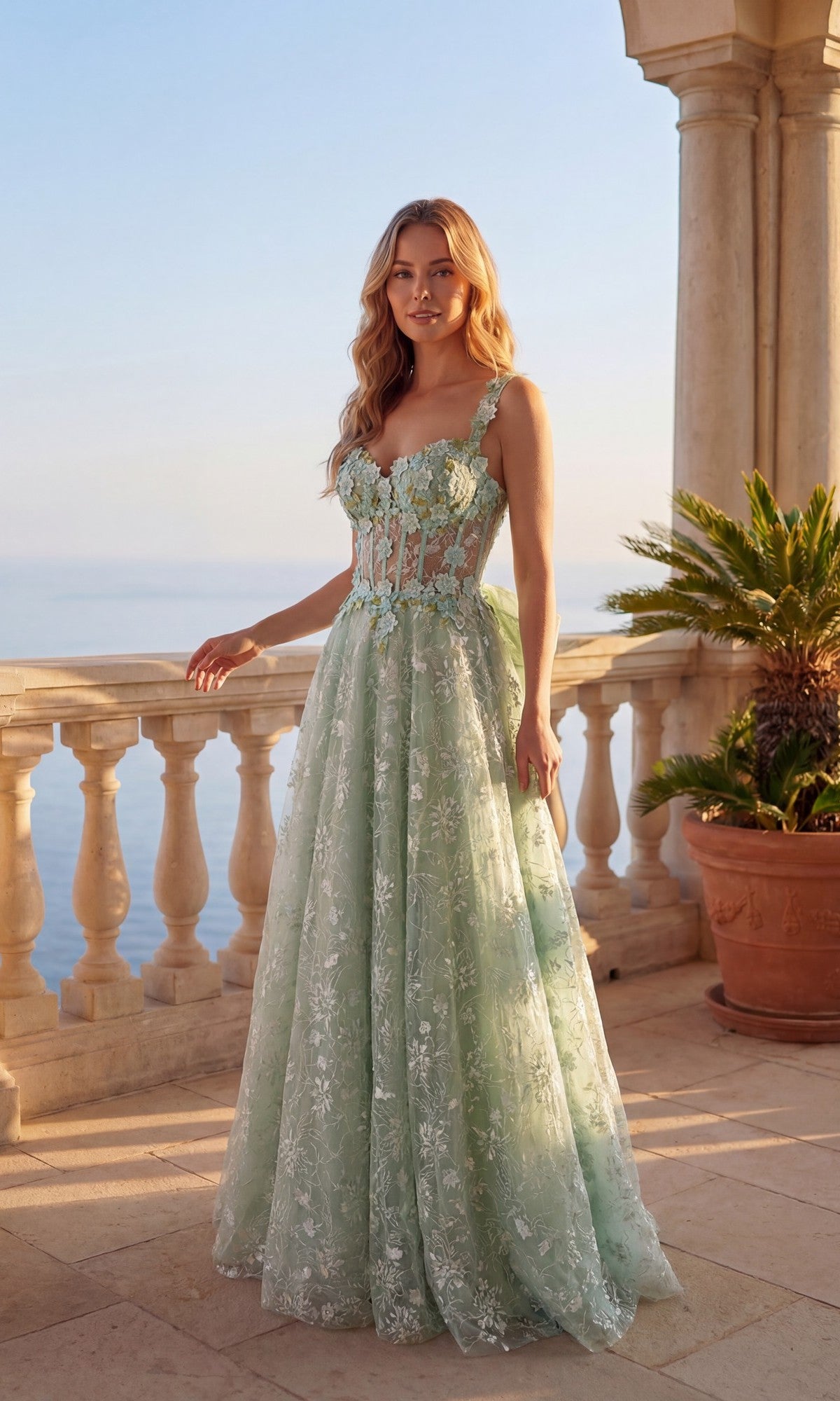 A woman in a sage Long Prom Dress: Chicas C370 with floral lace embroidery stands on a stone terrace, ocean and palm plants in the background.