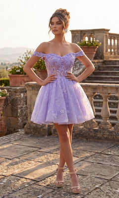 A woman wearing the Chicas C377 short homecoming dress in lilac, featuring an off-the-shoulder embroidered corset bodice and a delicate tulle skirt, stands outdoors on a stone terrace surrounded by greenery and potted plants.