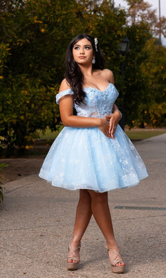 Wearing the "Short Homecoming Dress: Chicas C377" in sky blue, a woman stands outdoors on a pathway, the off-the-shoulder design and delicate tulle skirt flowing gracefully as lush greenery forms a picturesque backdrop.
