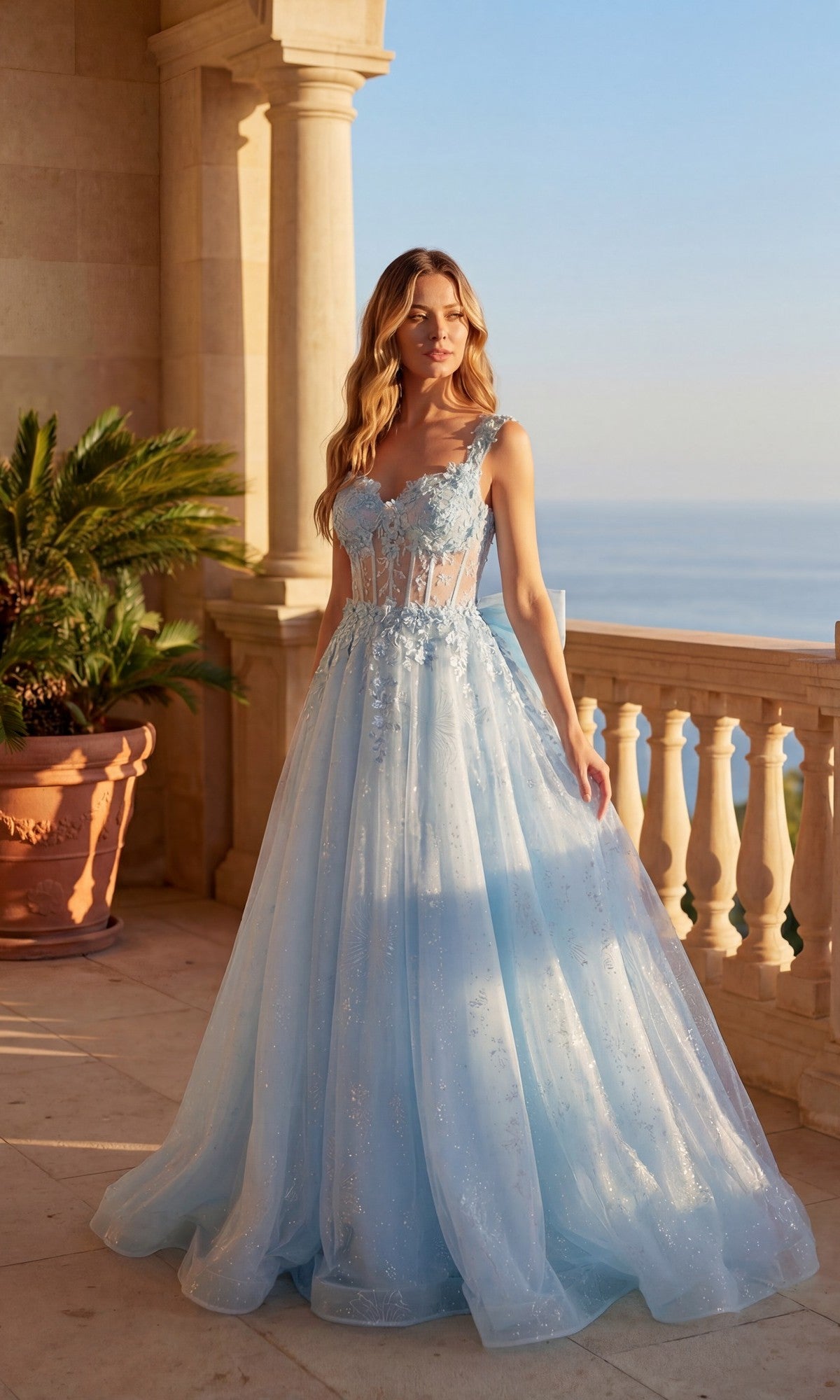 A woman in the sky blue Long Prom Dress: Chicas C379 with a flowing silhouette and floral details stands on a balcony overlooking the sea.