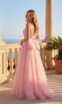 A woman in the Chicas C379 Long Prom Dress, a light pink A-line gown with a large back bow and floral sequin lace embroidery, stands on a stone terrace by the sea with a potted palm nearby.