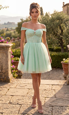 A woman stands outdoors in a sage Short Homecoming Dress: Chicas C382, featuring an off-the-shoulder sweetheart neckline, corset bodice, and tulle fit-and-flare skirt, paired with nude heels; garden scenery and a stone path are visible in the background.