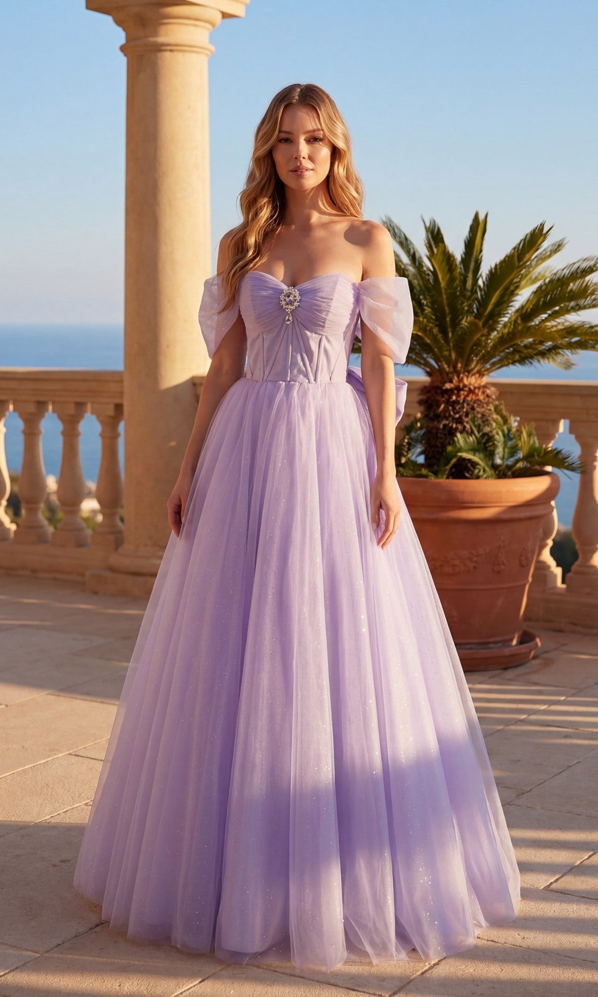 A woman poses outdoors by a stone balustrade and potted palm in the Chicas C386 Long Prom Dress, a floor-length, off-the-shoulder lavender gown with a tulle skirt, with the dreamy sea and sky as her backdrop.
