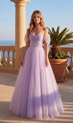 A woman poses outdoors by a stone balustrade and potted palm in the Chicas C386 Long Prom Dress, a floor-length, off-the-shoulder lavender gown with a tulle skirt, with the dreamy sea and sky as her backdrop.