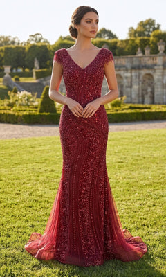 A woman stands on grass wearing the Chicas C389 long formal dress in burgundy, featuring a fitted, floor-length embellished design with beaded details, cap sleeves, and a V-neckline. Stone architecture and greenery appear in the background.