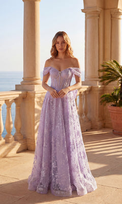 A woman in the Chicas C391 Long Prom Dress, a lilac off-the-shoulder gown with a sheer corset bodice and floral details, stands on a sunlit terrace overlooking the sea.