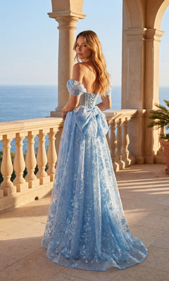 A woman in the Chicas C391 Long Prom Dress—light blue, off-the-shoulder with a floral glitter print and removable back bow—stands on a stone balcony by the sea as sunlight highlights her wavy hair.