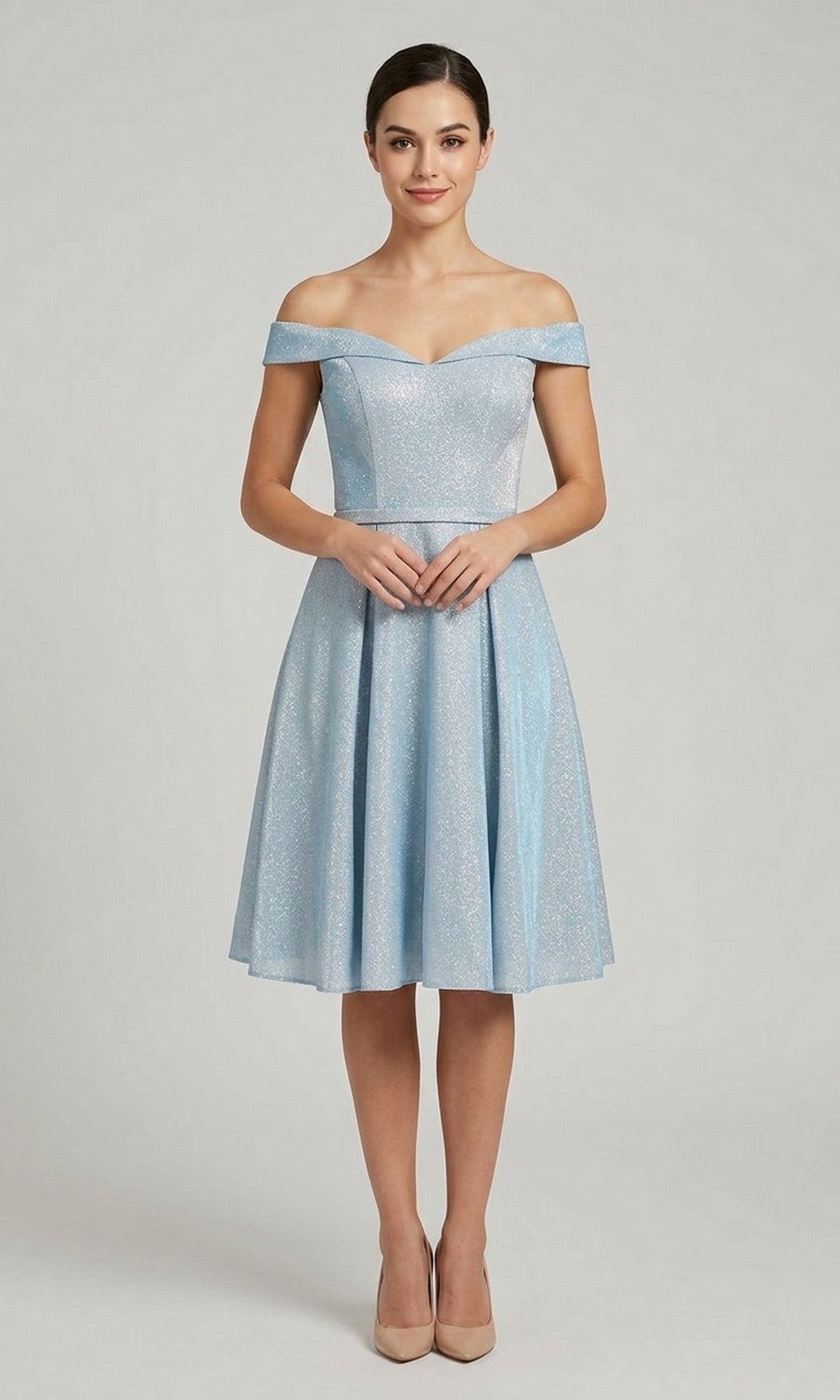 A person standing in a studio is wearing the Chicas C7518 Short Homecoming Dress in light blue, featuring a knee-length, sparkly design with an off-the-shoulder neckline and an elegant A-line skirt, paired with beige heels and hands clasped in front.