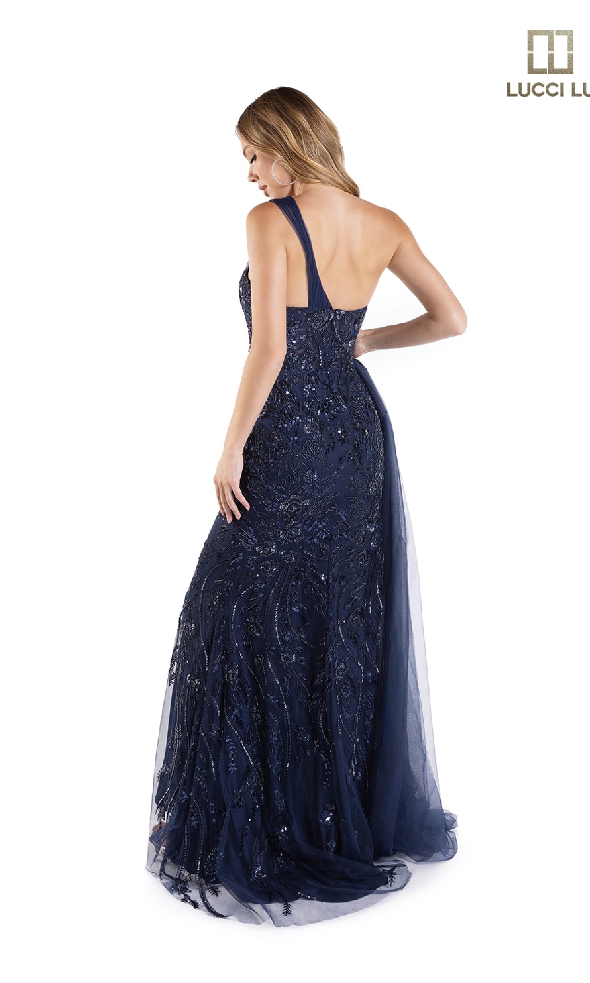 A woman stands facing away in the Lucci Lu C8041, a long navy blue prom dress featuring a one-shoulder design, floor-length silhouette, sequin embellishments, and a tulle side sash against a white background.
