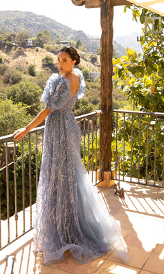 Wearing the Lucci Lu C8042 Long Prom Dress in blue, a woman with ruffled sleeves and a semi-sheer bodice stands on a sunlit balcony overlooking scenic hills.