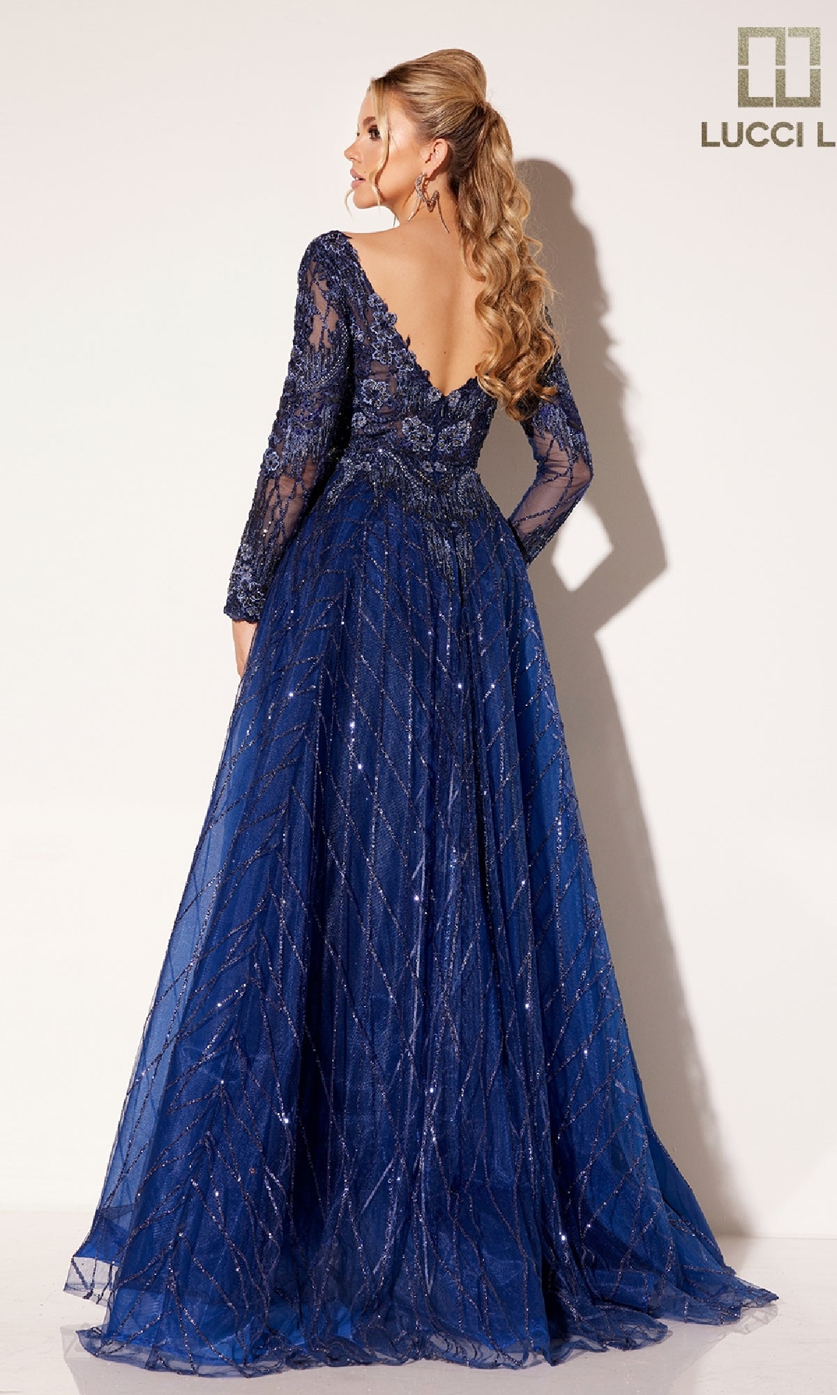 A woman with long wavy hair models the Lucci Lu C8076, a floor-length navy blue prom dress with long sleeves, lace details, and a plunging v-neckline, standing against a plain background.