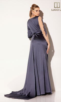A woman wearing the Lucci Lu C8104 long formal dress, in dark purple with sheer embroidered sleeves, a belt, and a floor-length train, stands facing a white wall while looking over her shoulder.