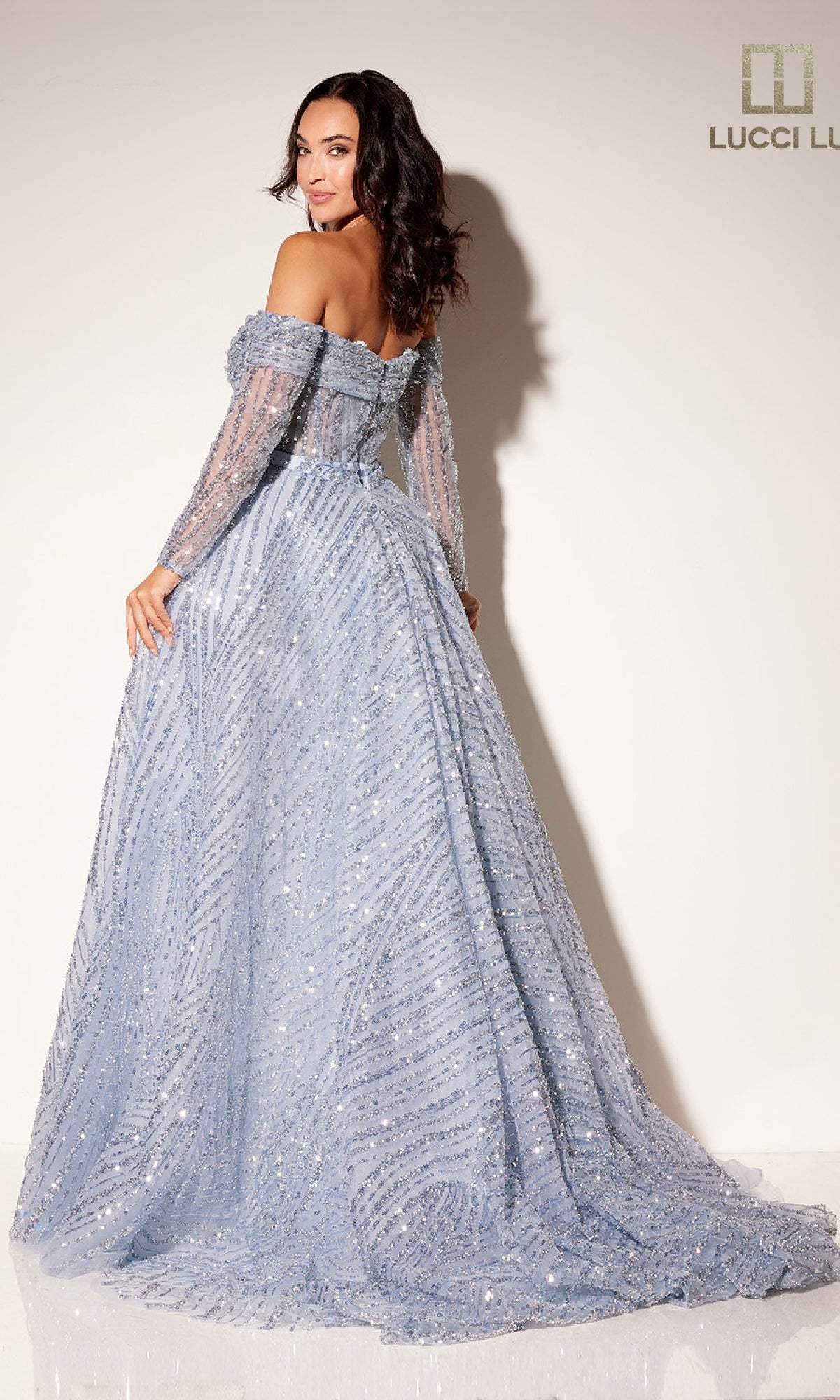 A woman poses with her back to the camera in the Lucci Lu C8112 long formal dress, featuring light blue sequins, an off-the-shoulder bodice, and long sleeves against a plain white wall.