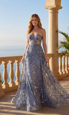 A woman in the smokey blue Chicas C819 long prom dress with silver embellishments and a sheer corset bodice stands on a sunlit terrace overlooking the sea.