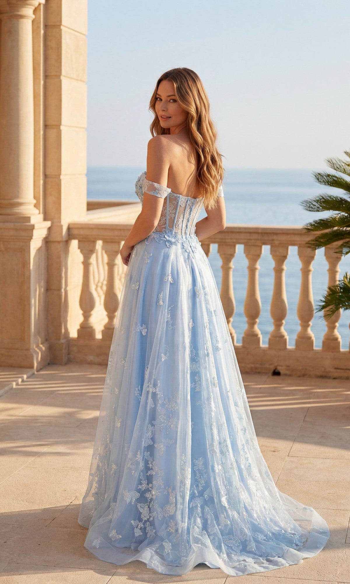 A woman in the Long Prom Dress: Chicas C822, a light blue off-the-shoulder gown with floral details, stands on a stone terrace overlooking the ocean, framed by palm leaves and ornate pillars.