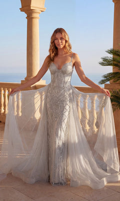 A woman in the Long Prom Dress: Chicas C824 in Silver/Silver, featuring beaded details, sheer panels, and a tulle overskirt, stands on a sunlit terrace with stone columns overlooking the sea.