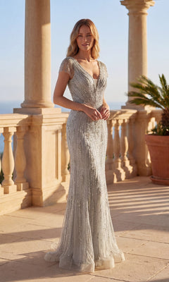 Wearing the Chicas C832 long formal dress in silver, a woman with sheer cap sleeves and beaded details stands on a sunlit terrace framed by stone columns and a potted palm.