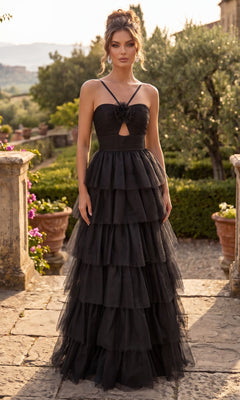 A woman stands outdoors in the Chicas C837 Long Prom Dress in black, featuring a layered tulle design with a floral detail at the bust and a dramatic tiered full skirt, surrounded by lush greenery and classic stone pathways.