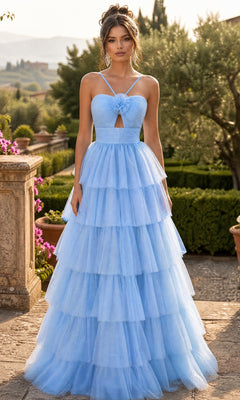 A woman stands outdoors surrounded by greenery and stone architecture, wearing the Chicas C837 Long Prom Dress in blue—a tiered tulle gown featuring thin straps, a corset back, and a keyhole cutout.