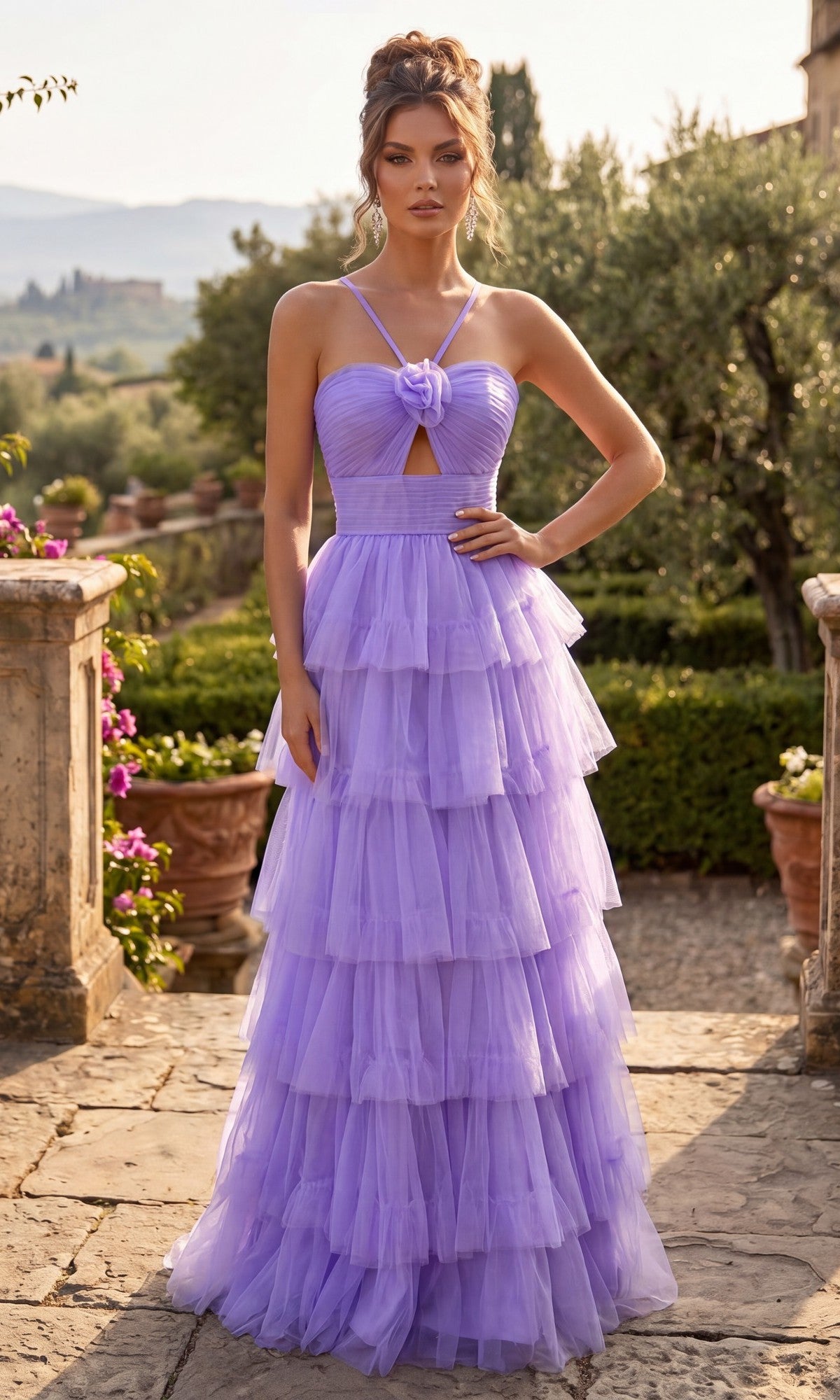 A woman stands outdoors on a stone terrace, wearing the Chicas C837 Long Prom Dress in lavender—a layered tulle ball gown with a corset back and spaghetti straps. Her tiered full skirt flows gracefully amid lush greenery and distant hills in the background.