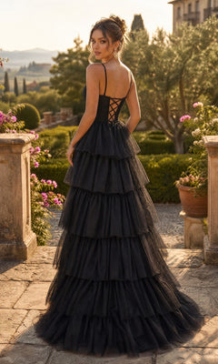 Wearing the Long Prom Dress: Chicas C837, a woman stands outdoors amidst lush greenery and stone architecture. The tiered black evening gown features a corset back and thin straps, with its layered tulle full skirt cascading elegantly around her.