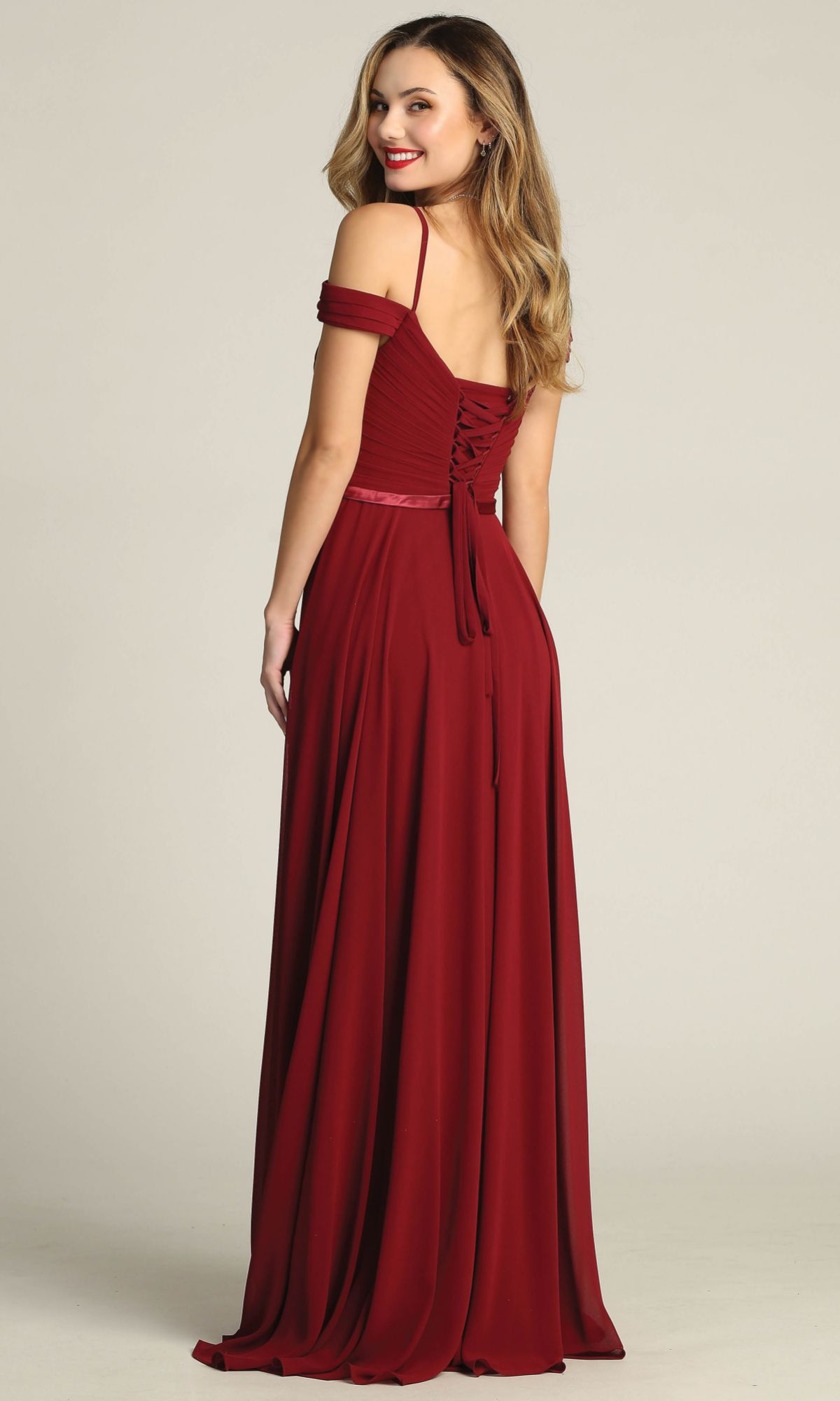 Wearing the Chicas C8997 Long A-Line Off-Shoulder Prom Dress featuring a fitted bodice and lace-up back, a woman stands facing away and smiles over her shoulder.
