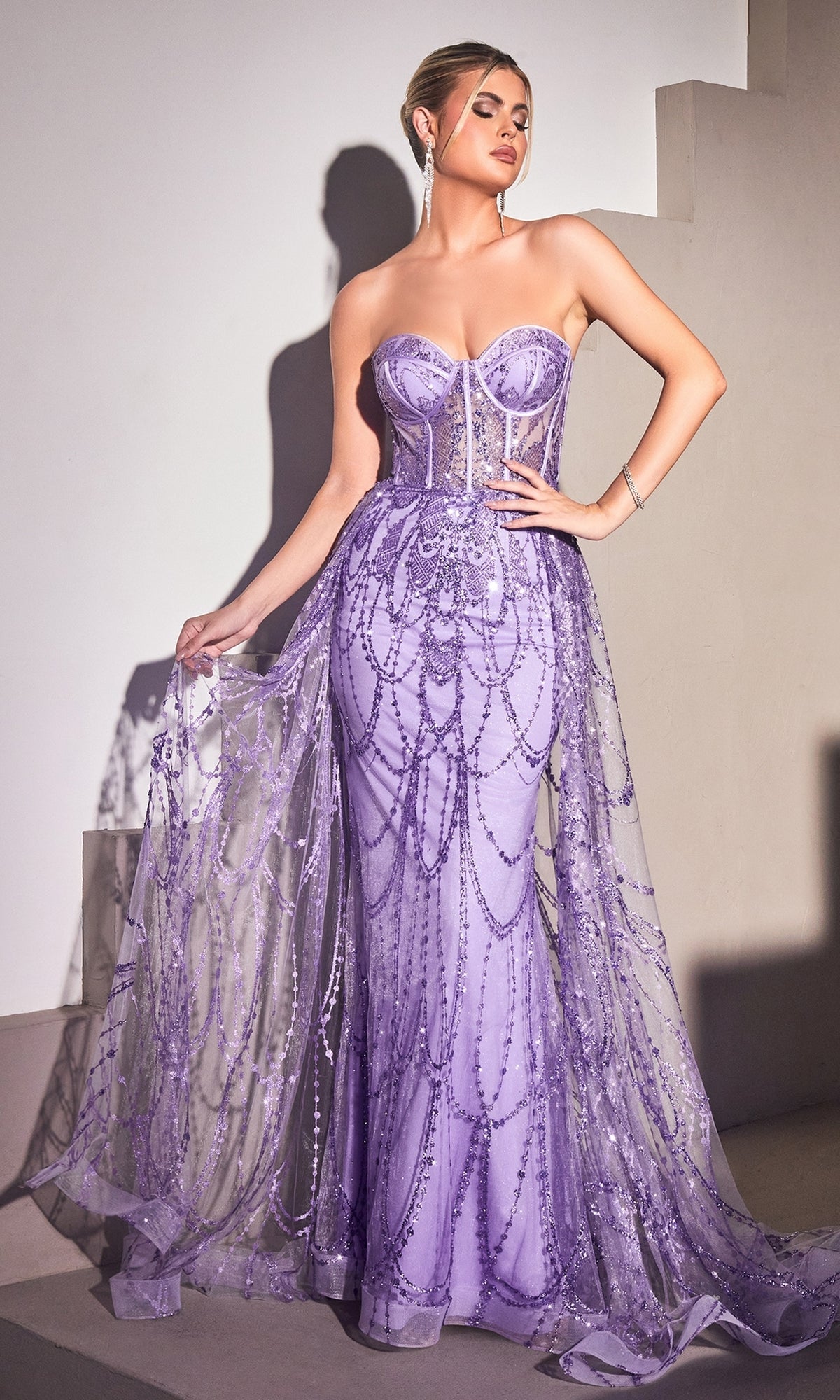 A woman stands elegantly on a staircase wearing the Strapless Mermaid Prom Dress with Overskirt CB095, featuring a lilac hue, fitted bodice, beaded details, and a sheer flowing overlay.