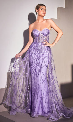 A woman stands elegantly on a staircase wearing the Strapless Mermaid Prom Dress with Overskirt CB095, featuring a lilac hue, fitted bodice, beaded details, and a sheer flowing overlay.
