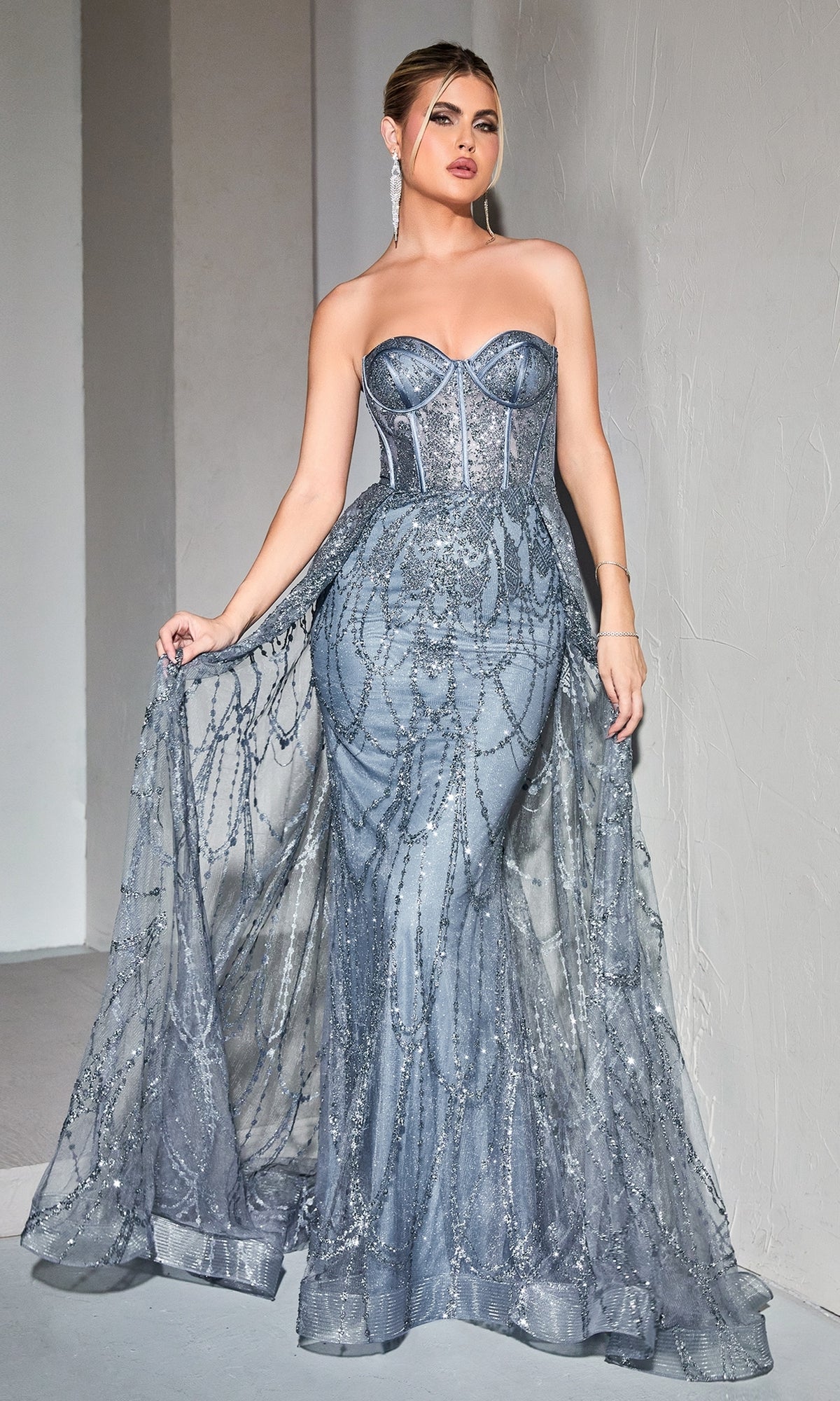 A woman poses in the Strapless Mermaid Prom Dress with Overskirt CB095, a formal silver-blue gown with sheer embellished overlay, standing against a light textured wall.
