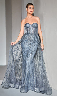 A woman poses in the Strapless Mermaid Prom Dress with Overskirt CB095, a formal silver-blue gown with sheer embellished overlay, standing against a light textured wall.