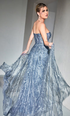 Woman wearing the Strapless Mermaid Prom Dress with Overskirt CB095, featuring blue-gray sequins, holds out the skirt and looks over her shoulder in a softly lit room.