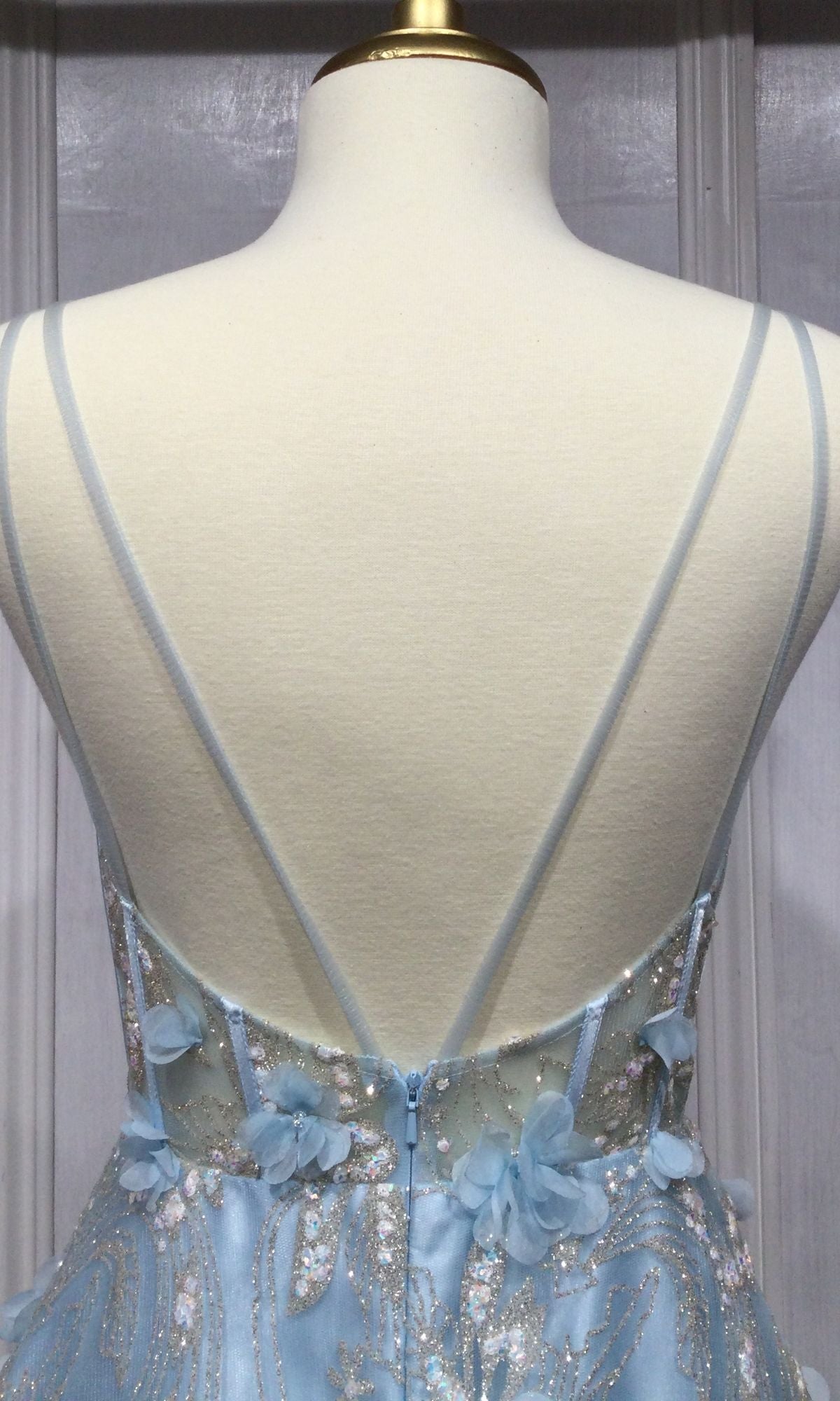 The Ladivine Long Formal Dress CB105 is shown on a mannequin, featuring a light blue ball gown with thin crisscross straps, 3D floral accents, silver embroidery, and a sheer corset bodice.