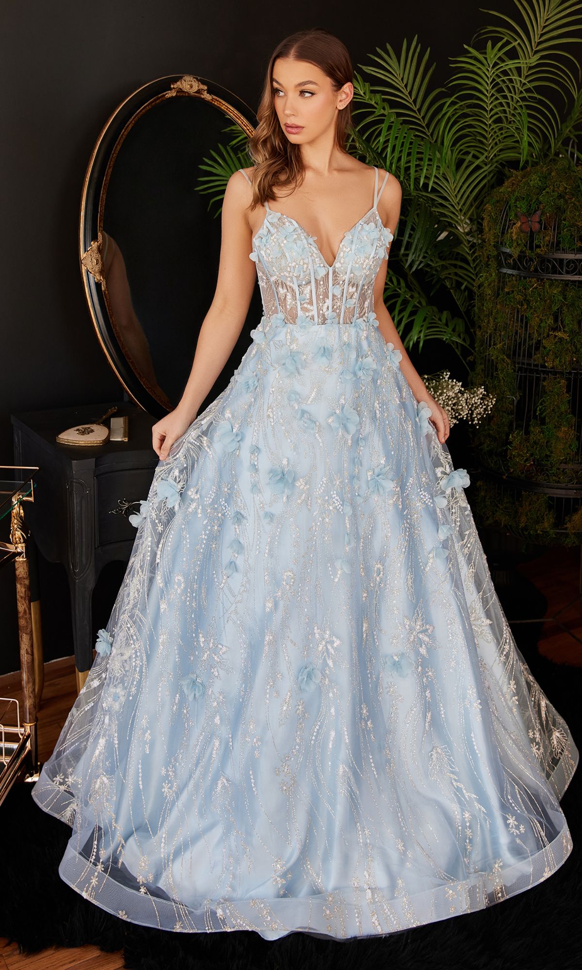 A woman wears the Ladivine Long Formal Dress CB105, featuring a blue ball gown design with 3D floral details and a sheer corset bodice, standing indoors beside a mirror and lush green plants.