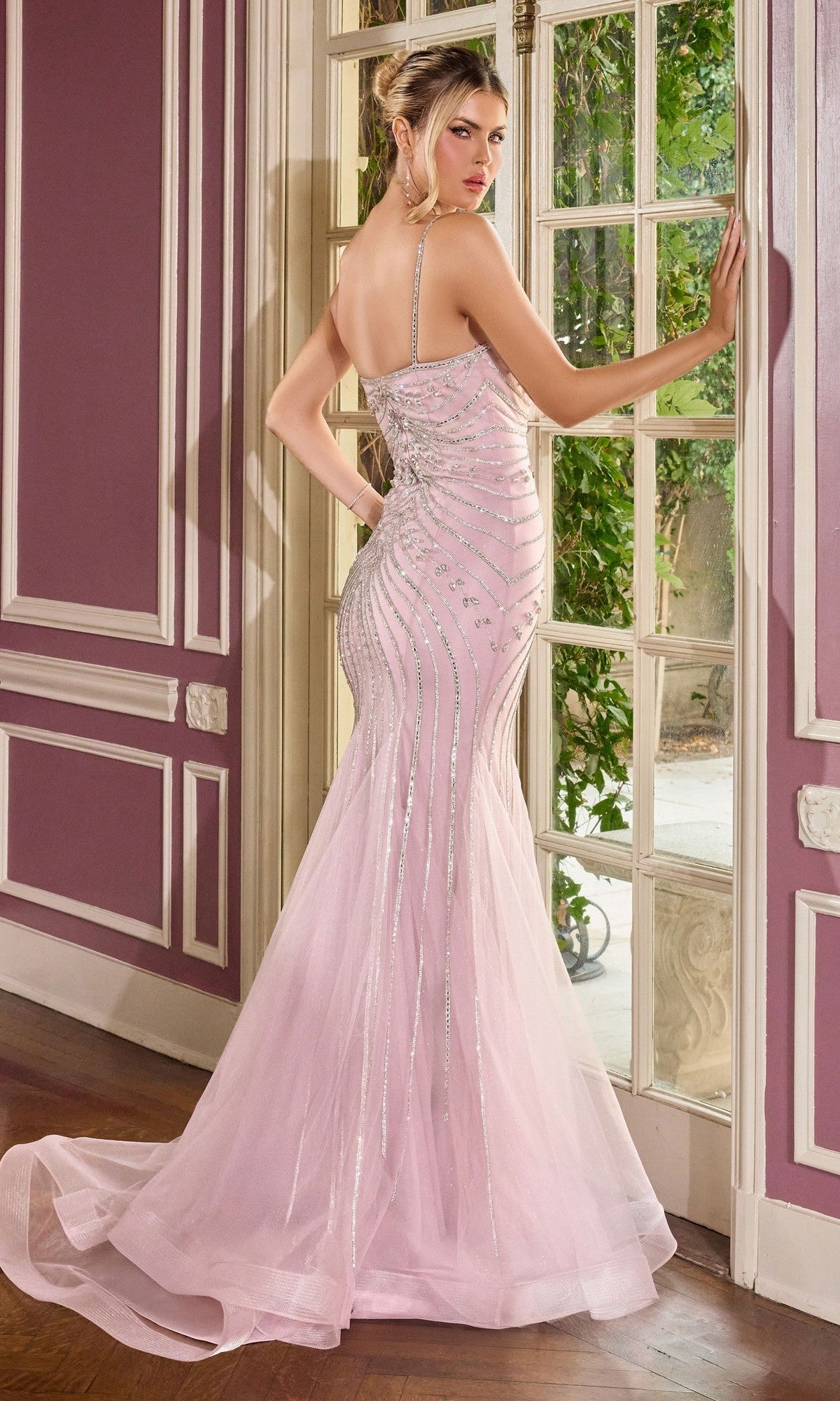 A woman wearing the Ladivine CB167 long prom dress—a pale pink, form-fitting, floor-length mermaid gown with silver embellishments—stands by a large window, glancing over her shoulder.