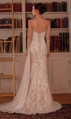 A woman in the Ladivine CB168W Long Prom Dress, a strapless gown adorned with delicate floral lace appliques and featuring a sheer train, stands in front of a bookcase, facing away from the camera.