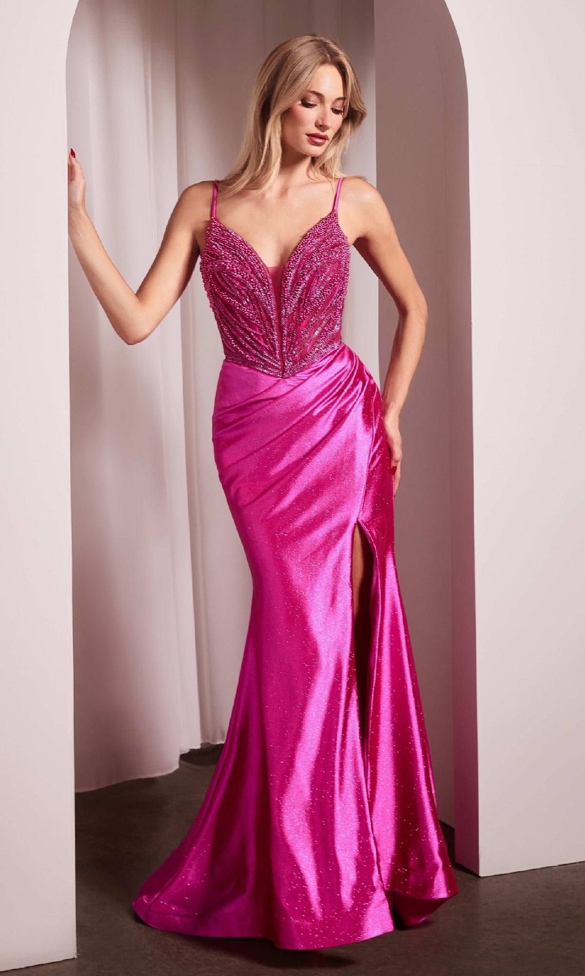 A woman stands in an archway wearing the Ladivine CC0238 Beaded-Bodice Long Prom Dress, a bright pink gown with pearlized beading on the bodice, thin straps, and a thigh-high slit.