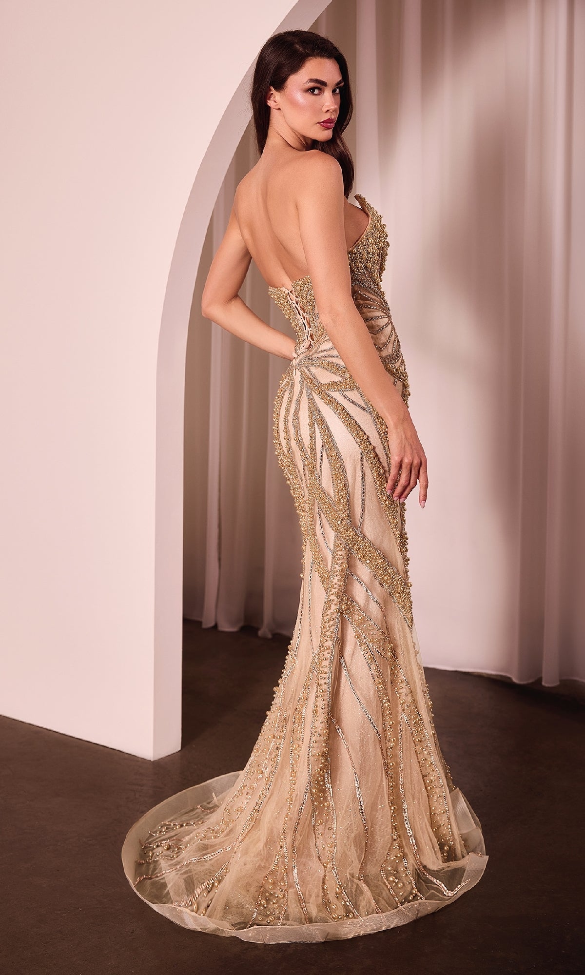 A woman wearing the Ladivine CC0688 Strapless Long Beaded Prom Dress with a corset bodice stands in front of a white curtain, looking over her shoulder.
