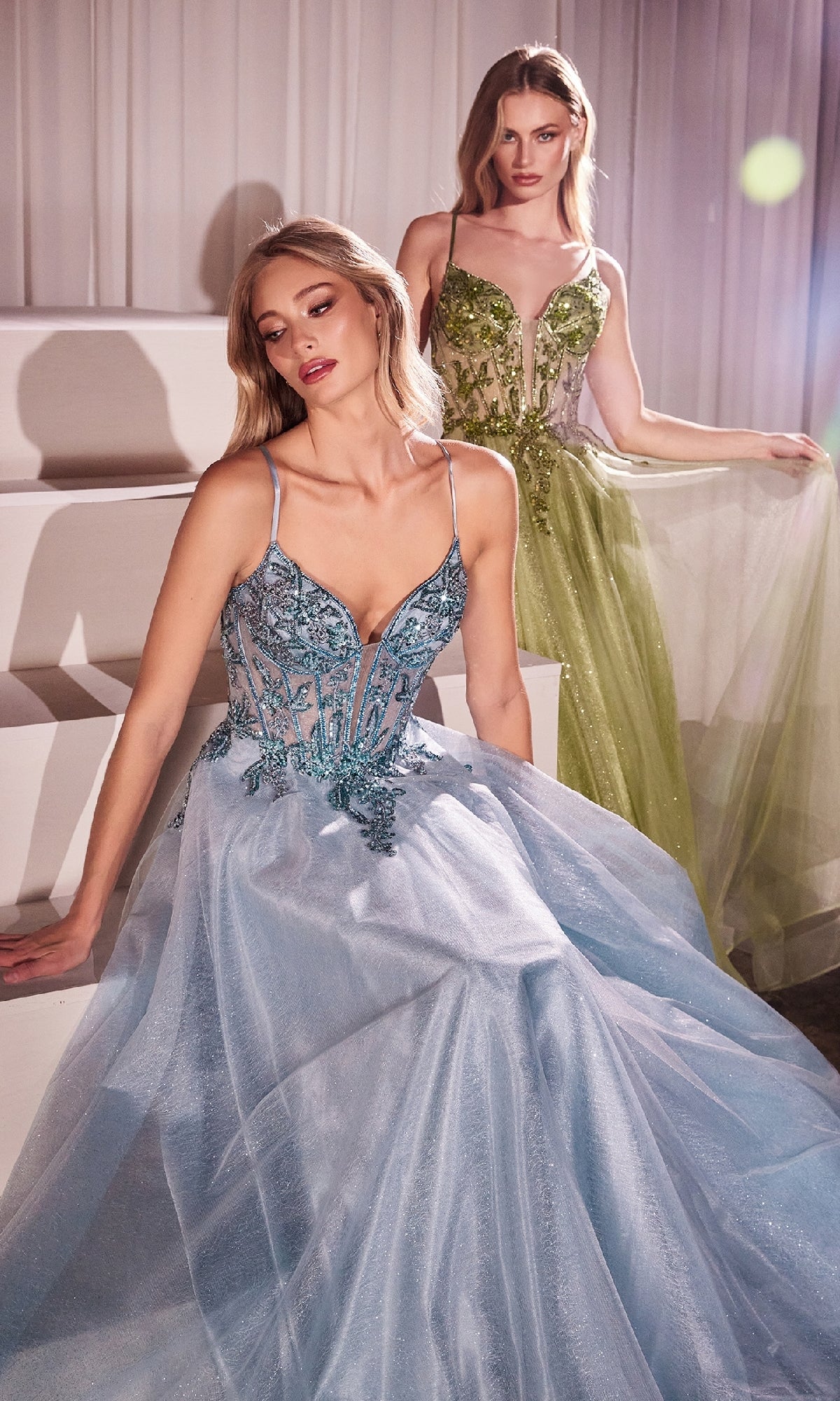 Two women pose indoors in floor-length evening gowns: one wears a light blue formal dress with floral details, the other dons the Sheer-Corset Long Prom Dress: Ladivine CC0881, a green gown with beaded embroidery and sheer accents.