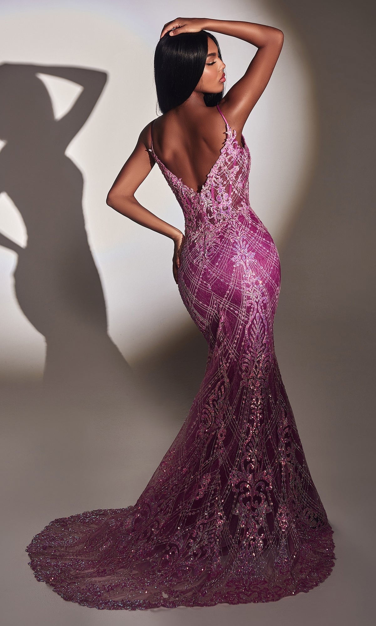 A woman poses with one hand on her head in the Amethyst Long Sequin Mermaid Prom Dress CC2168, a fitted, floor-length gown with a deep V-back, against a neutral background with her shadow behind her.