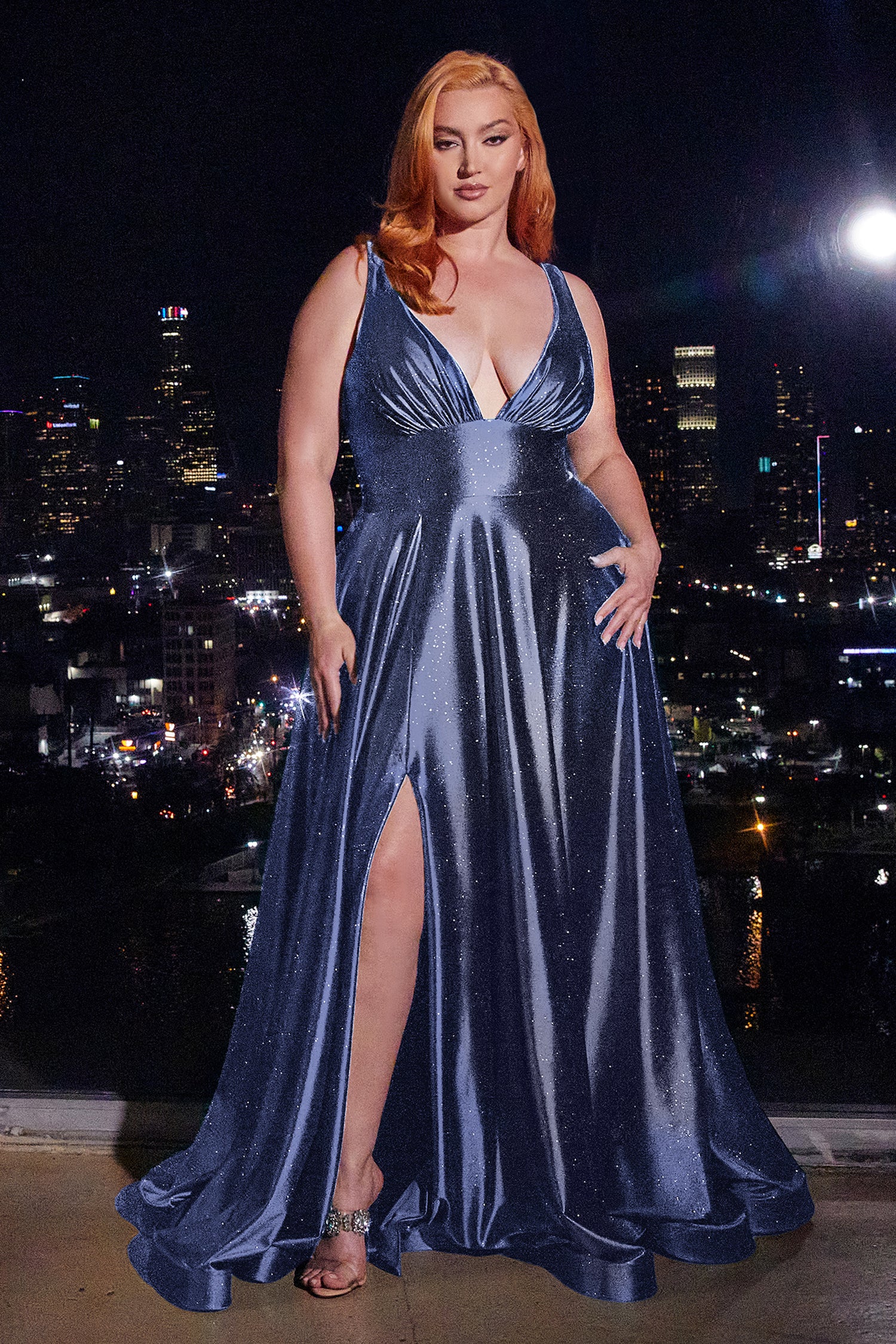 A woman with red hair in the Ladivine CC2349C Plus Long Glitter Prom Dress stands on a balcony at night, city lights sparkling in the background.