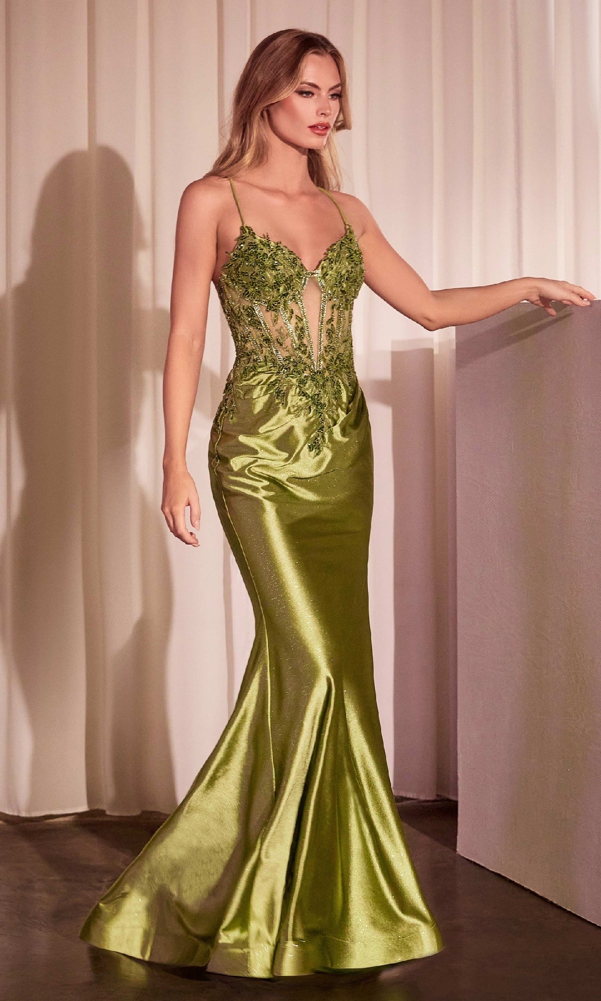 A woman stands indoors in the Ladivine CC2401 Lace-Up Long Corset Prom Dress, a floor-length, shiny olive green gown with a beaded corset bodice, floral lace details, and an elegant lace-up back.