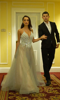 A woman in the Ladivine CC2531 long silver a-line prom dress with a beaded deep v-neck bodice walks hand in hand with a man in a black suit and white shirt down a hallway with yellow walls and red carpet.