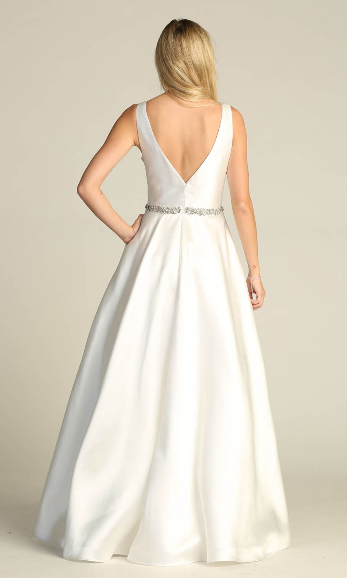 A woman stands with her back to the camera, wearing the Jeweled-Waist Long Prom Ball Gown Chicas CC3021—a sleeveless white A-line dress with an embellished waistband and a deep V-back.