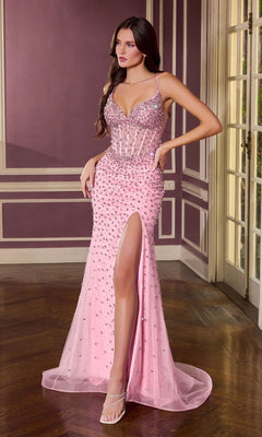 A woman wears the Ladivine CC3902 Long Prom Dress in pink, featuring crystal embellishments, a sheer corset bodice, thigh-high slit, and spaghetti straps as she stands in an elegant room with wood floors and panelled walls.
