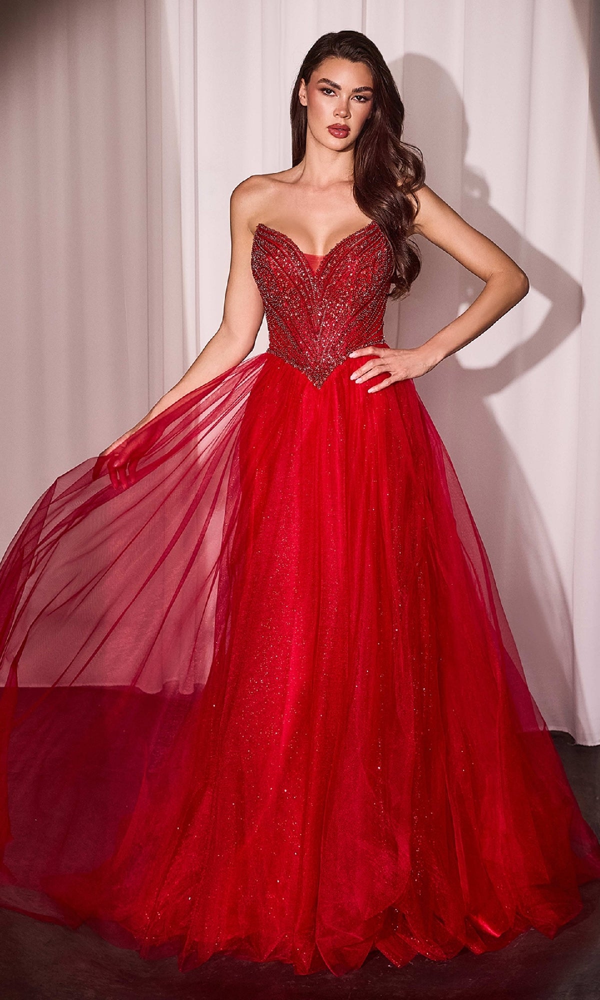 A woman wearing the Beaded-Bodice Long Prom Dress Ladivine CC415, featuring a fitted beaded bodice and flowing tulle skirt, stands in front of white curtains.