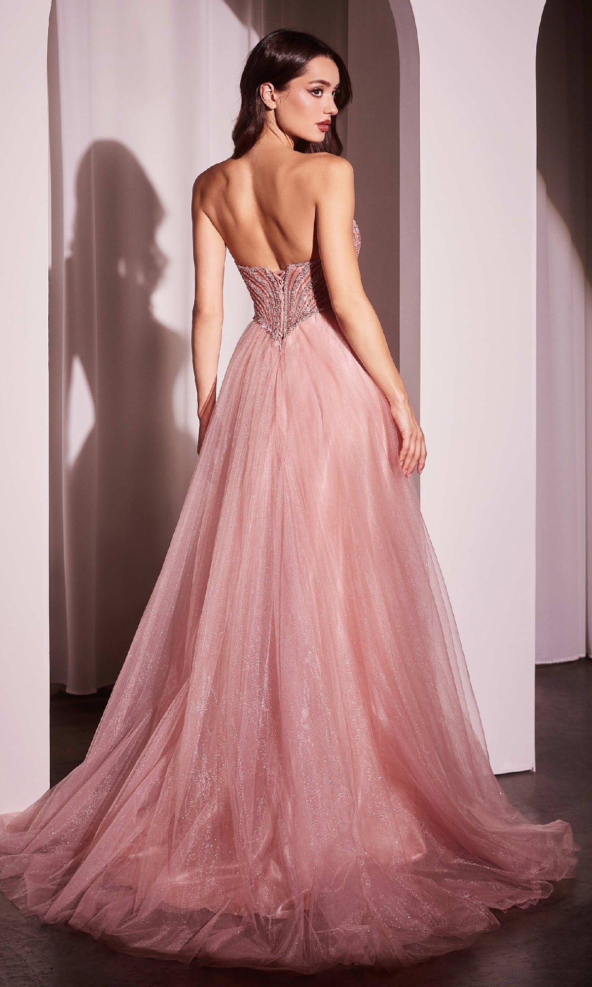 A woman in the Beaded-Bodice Long Prom Dress: Ladivine CC415, featuring a strapless fitted bodice and flowing tulle skirt, stands with her back to the camera in a softly lit room.