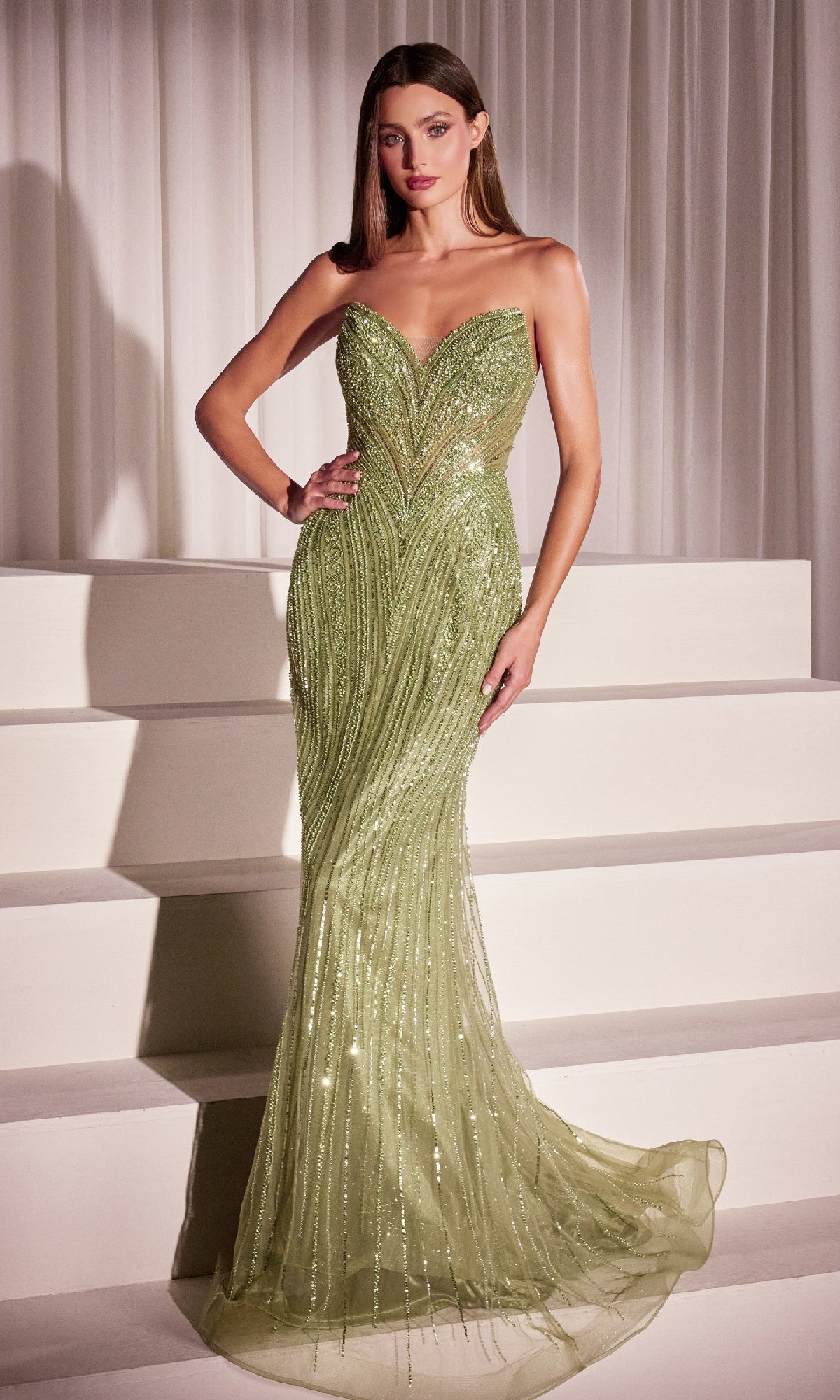A woman stands on white steps in the Ladivine CC416 Strapless Long Sequin Prom Dress in Greenery, a fitted evening gown adorned with shimmering sequins and beads, featuring a slight train and sleek straight hair.