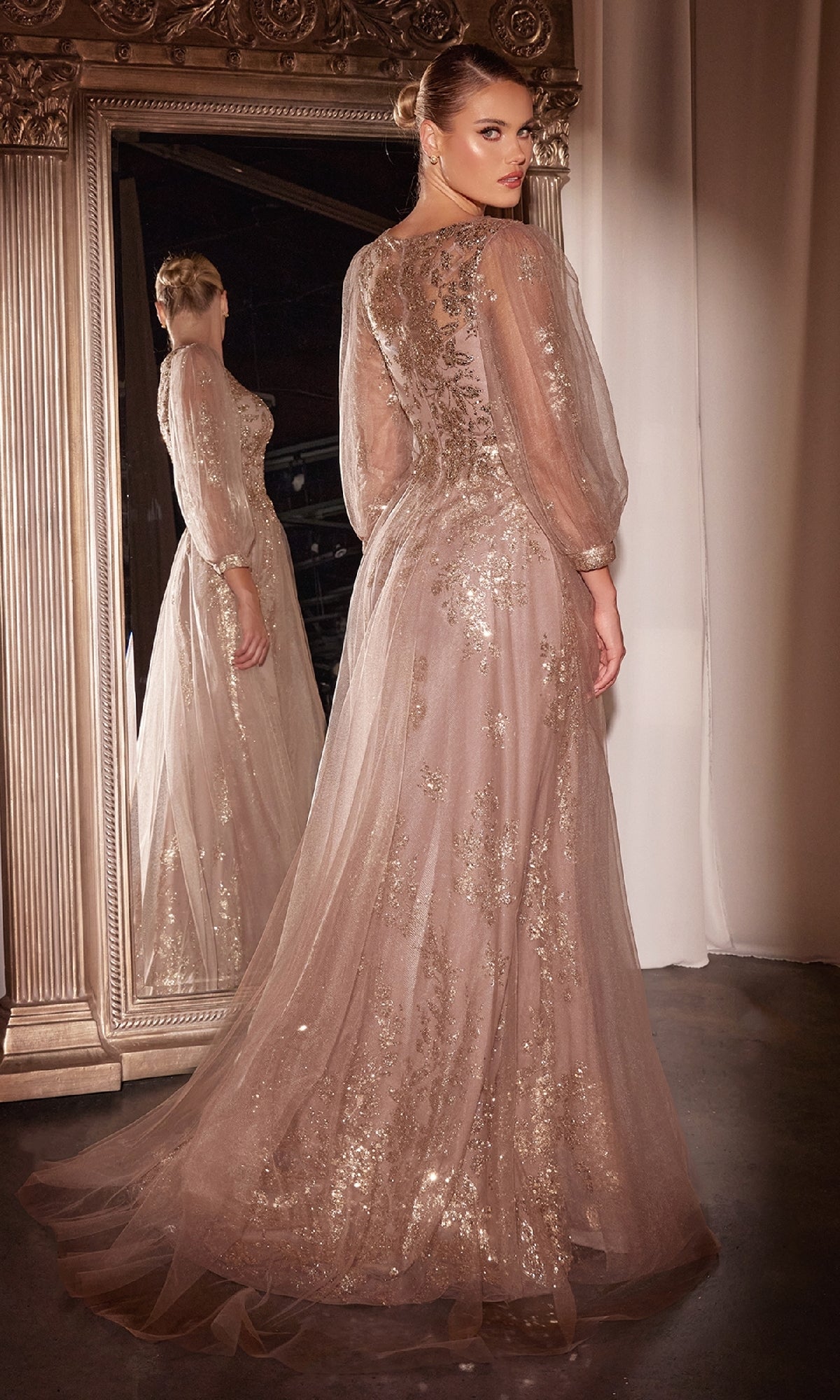 A woman in the Ladivine CD0102 Sheer-Sleeve Long Prom Dress with gold embellishments stands before a large ornate mirror, glancing back over her shoulder.